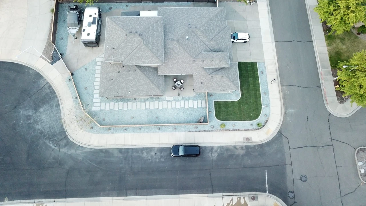 Overhead view of a residential corner lot with a house, driveway, light gravel border, a rectangular patch of green turf, parked vehicles, and surrounding streets and neighboring homes.