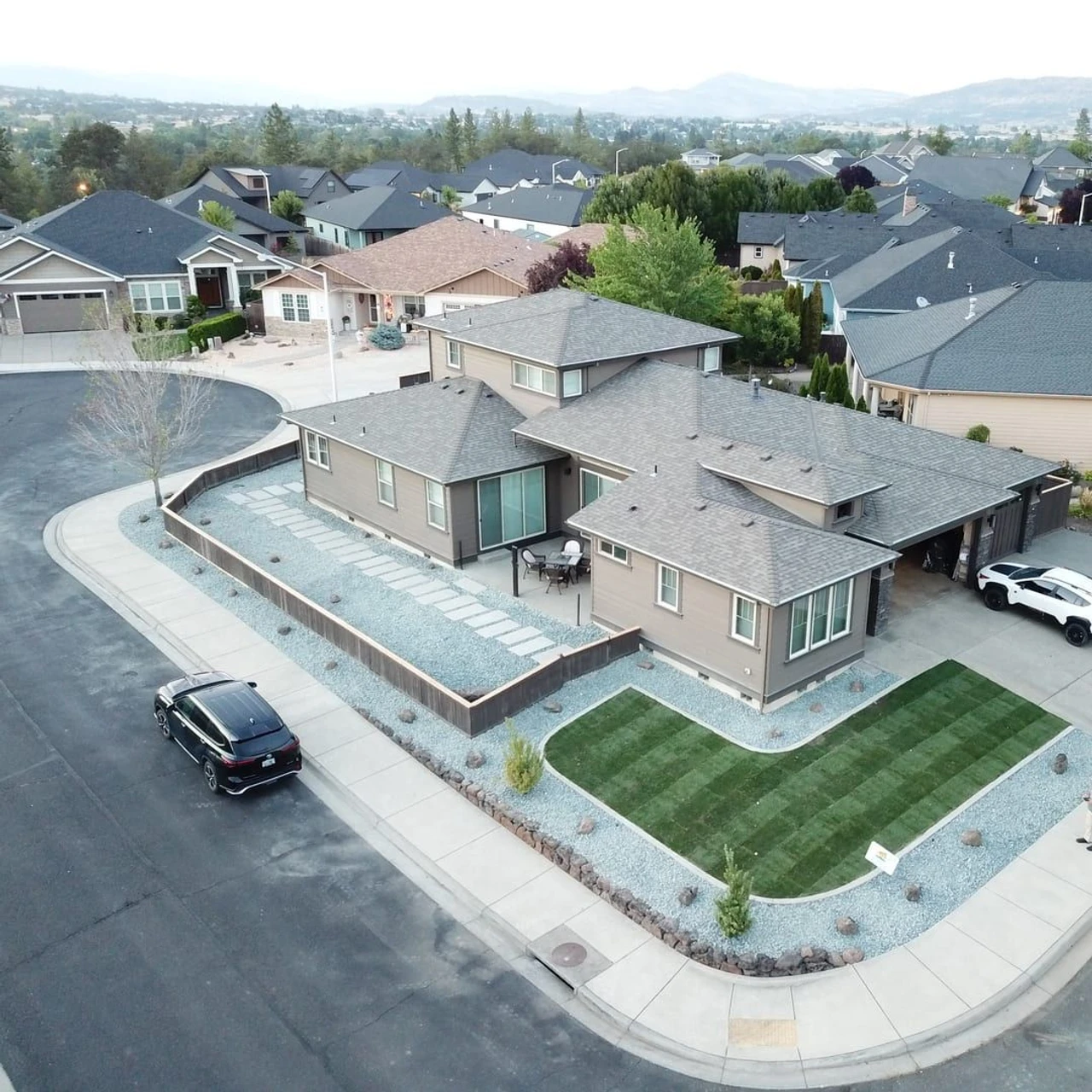 Aerial view of a suburban house with a newly landscaped front yard featuring a striped green lawn, gravel beds, stepping-stone paths, a concrete driveway, and parked cars on a residential street.