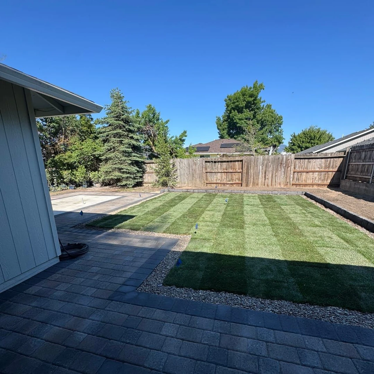 A backyard with a dark paver patio in the foreground, a striped synthetic turf lawn bordered by gravel, a wooden fence, trees, and a clear blue sky.