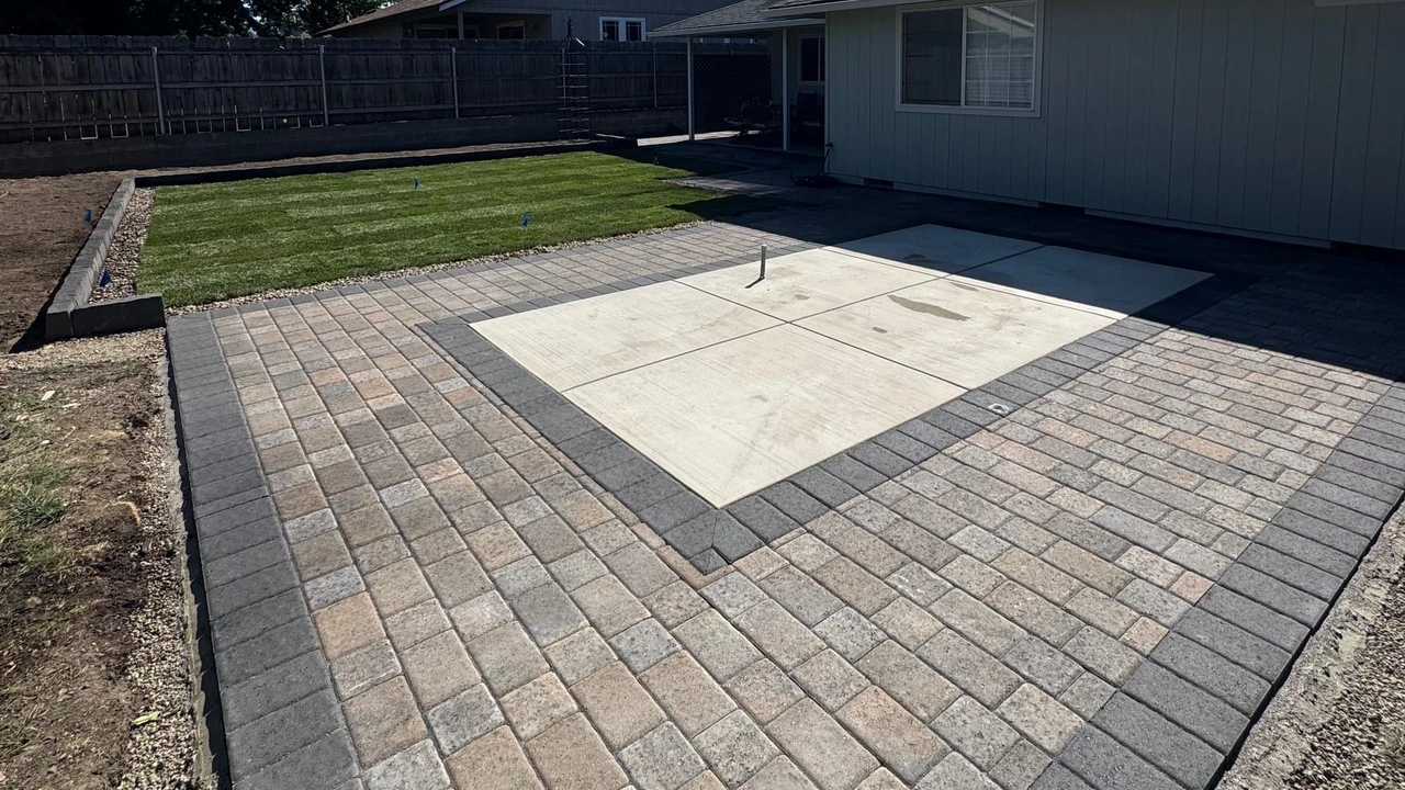 Backyard with a gray paver patio surrounding a square concrete slab, a small strip of grass lawn, a house wall, wooden fence, and tall trees under a bright sunny blue sky.