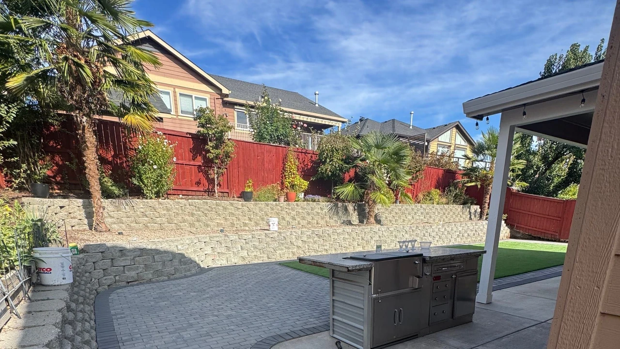 A backyard with a curved gray paver patio and walkway beside a concrete slab, stone retaining walls, a strip of artificial turf, potted plants, a red fence, and a covered outdoor kitchen under a blue sky.