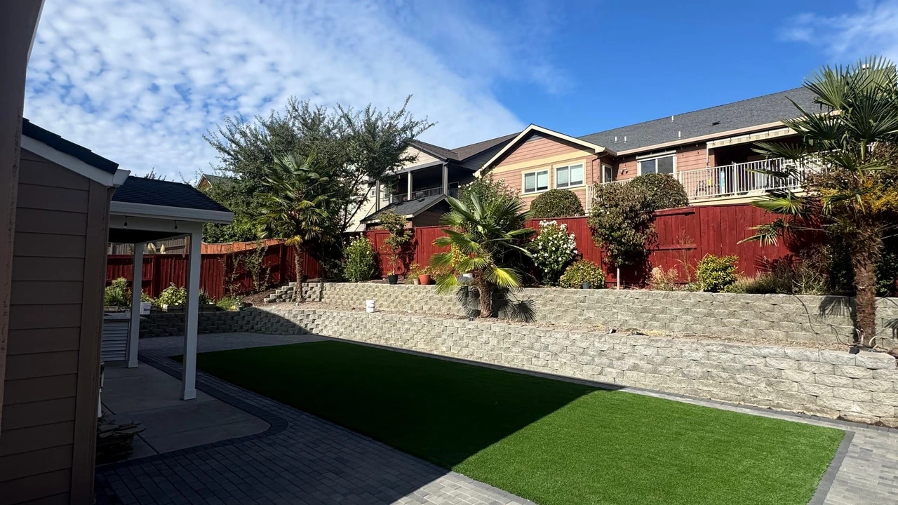Residential backyard with a gray paver patio, a rectangular synthetic turf lawn, a stacked stone retaining wall, a red fence, and several palm-like plants under a blue sky.