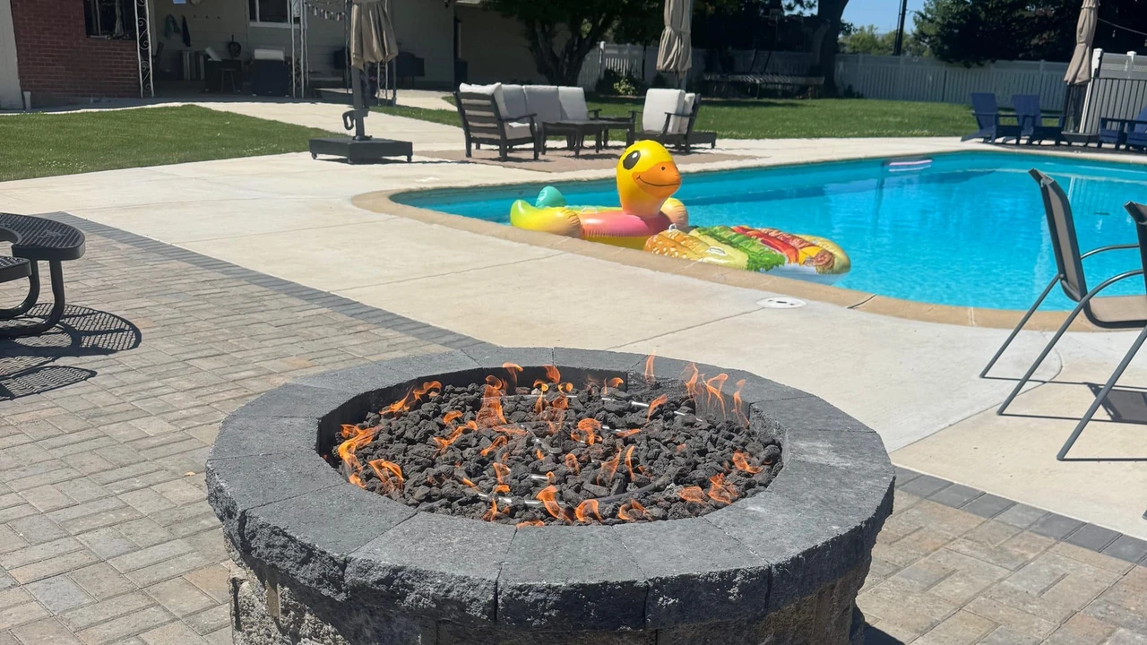 A stone fire pit with small flames sits on a paver patio beside a swimming pool, with patio chairs, umbrellas, a house, and large trees in the background.