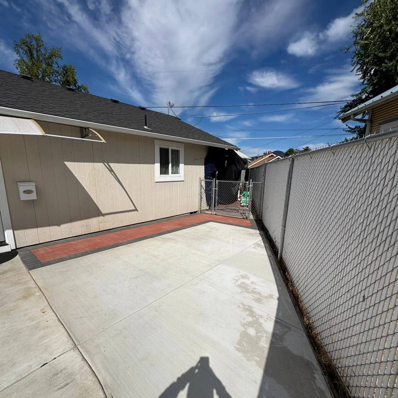 A residential side yard with a large concrete patio bordered by red brick, a beige house wall, a chain-link gate, and a white chain-link fence under a blue sky with clouds.