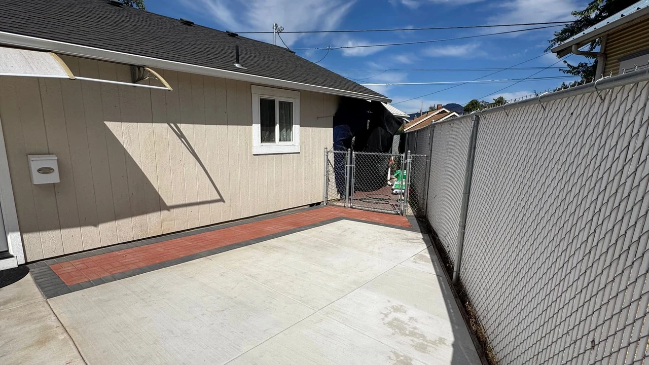 A residential side yard with a large concrete patio bordered by red brick, a beige house wall, a chain-link gate, and a white chain-link fence under a blue sky with clouds.