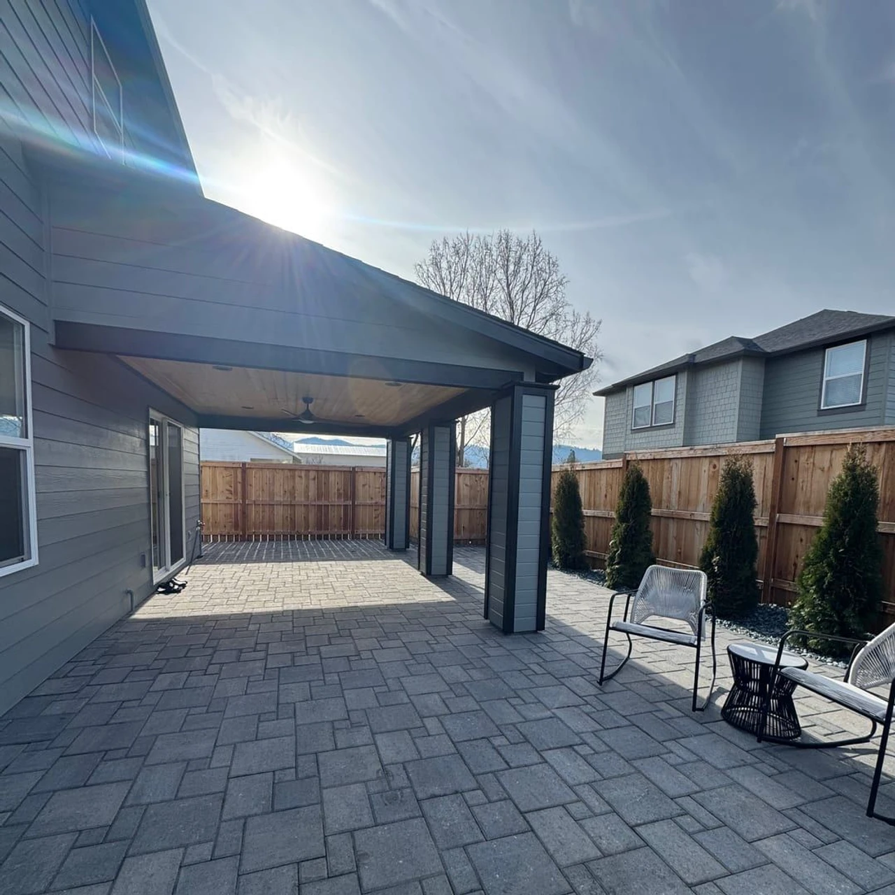 Gray paver patio beside a house under a covered roof with a ceiling fan, bordered by a wooden fence and evergreen shrubs. Two outdoor chairs and a small table sit near the fence in bright sunlight.