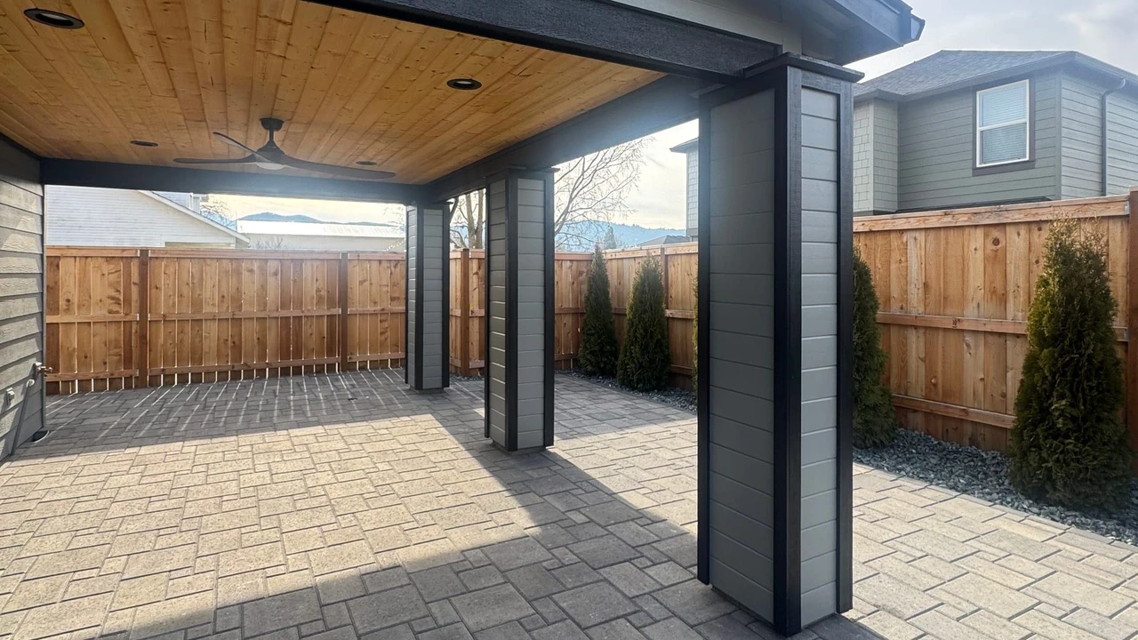 Covered backyard patio with gray paver flooring, dark support posts, wood-paneled ceiling, a wooden fence, and small evergreen shrubs along the perimeter.