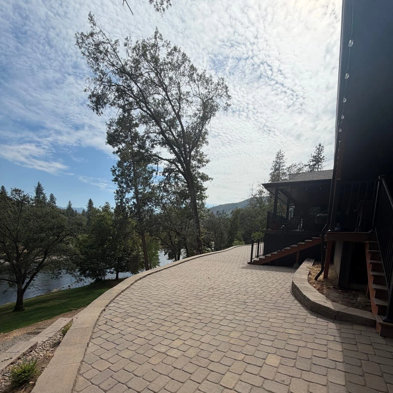 A curved stone paver driveway or patio beside a house, with stairs and a deck on the right, trees and a river in the background, and a cloudy sky above.