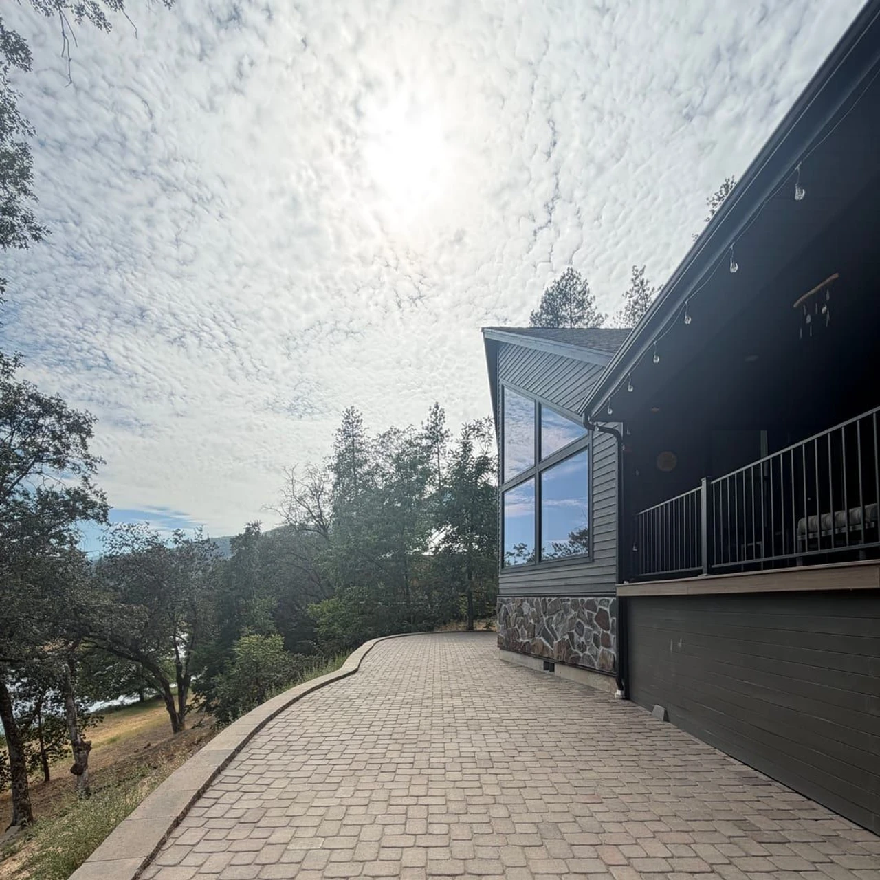 A curved paver walkway runs beside a house with a raised porch, stone foundation, and dark siding, overlooking trees and a hillside under a bright cloudy sky.