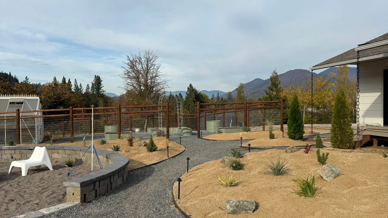 A landscaped backyard with a curved gravel path, dry planting beds, small shrubs and rocks, low path lights, a fenced perimeter, a house edge, and mountains under a cloudy sky.