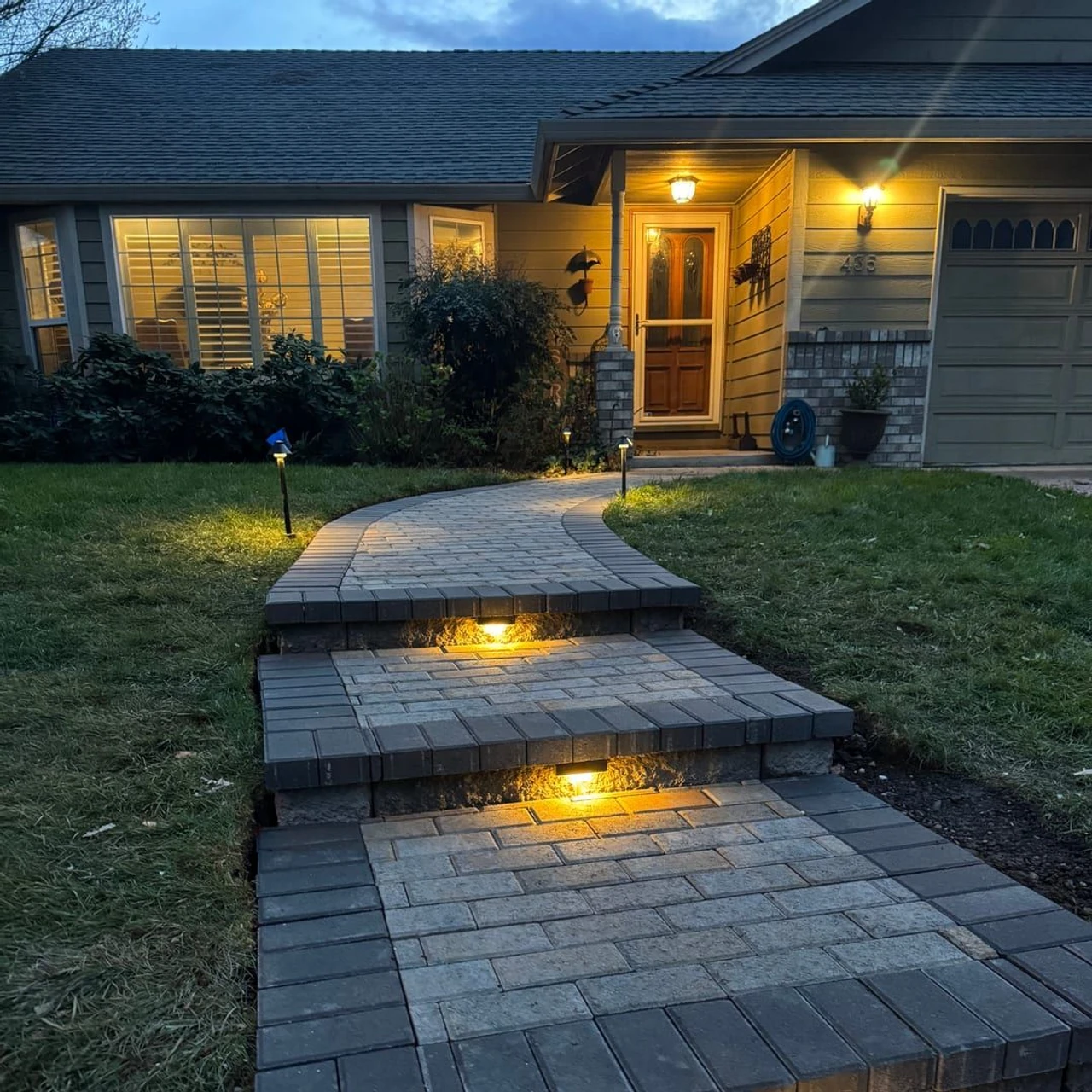 Curved paver front walkway with three raised steps and small встроенные lights leading to a house entrance at dusk, bordered by grass and shrubs, with warm porch and garage lights on.