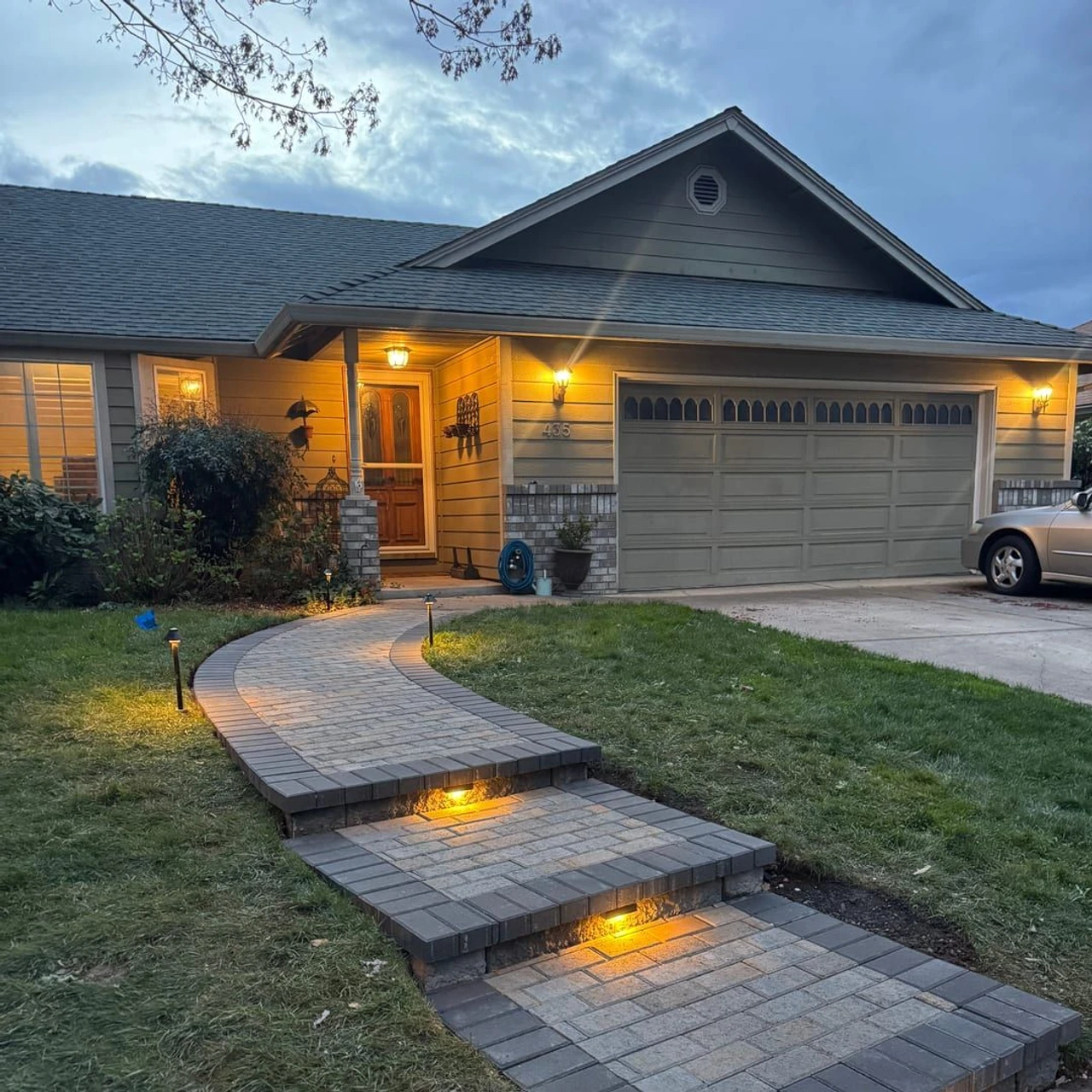 Curved paver walkway with short steps and warm ground lights leading to a house entrance and garage at dusk, with trimmed grass on both sides.