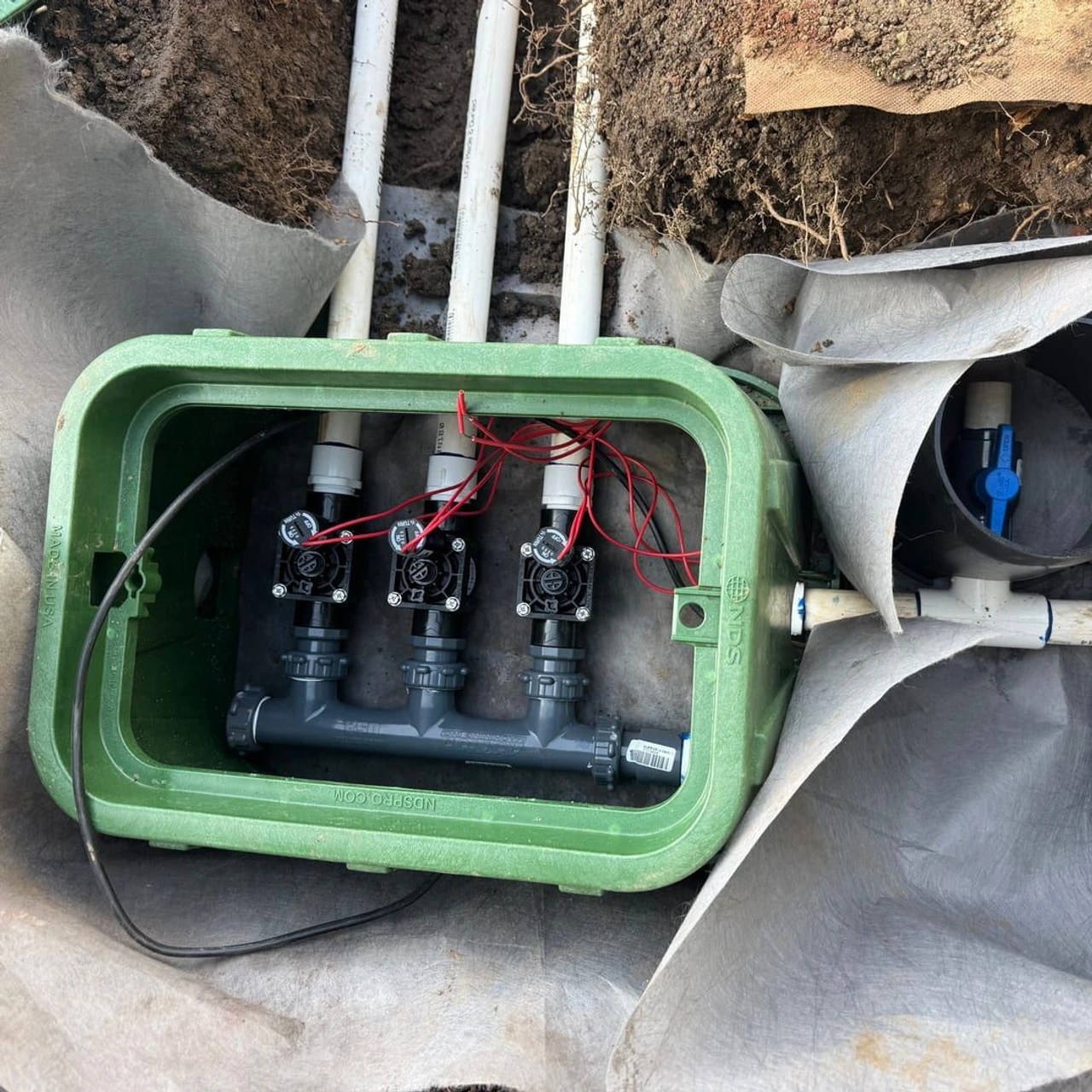 An open green underground valve box in dirt with three black irrigation valves, white PVC pipes, red wires, and landscape fabric folded around the excavation.