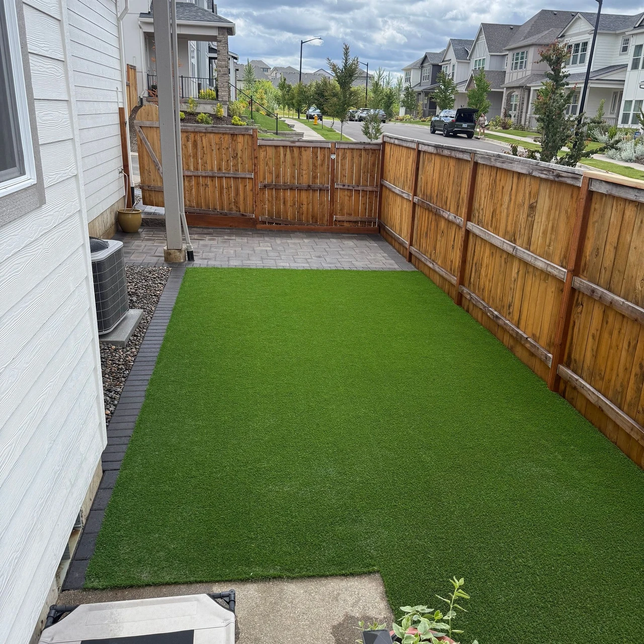 A white house to the left, wooden fence to the right. Backyard contains green artificial turf