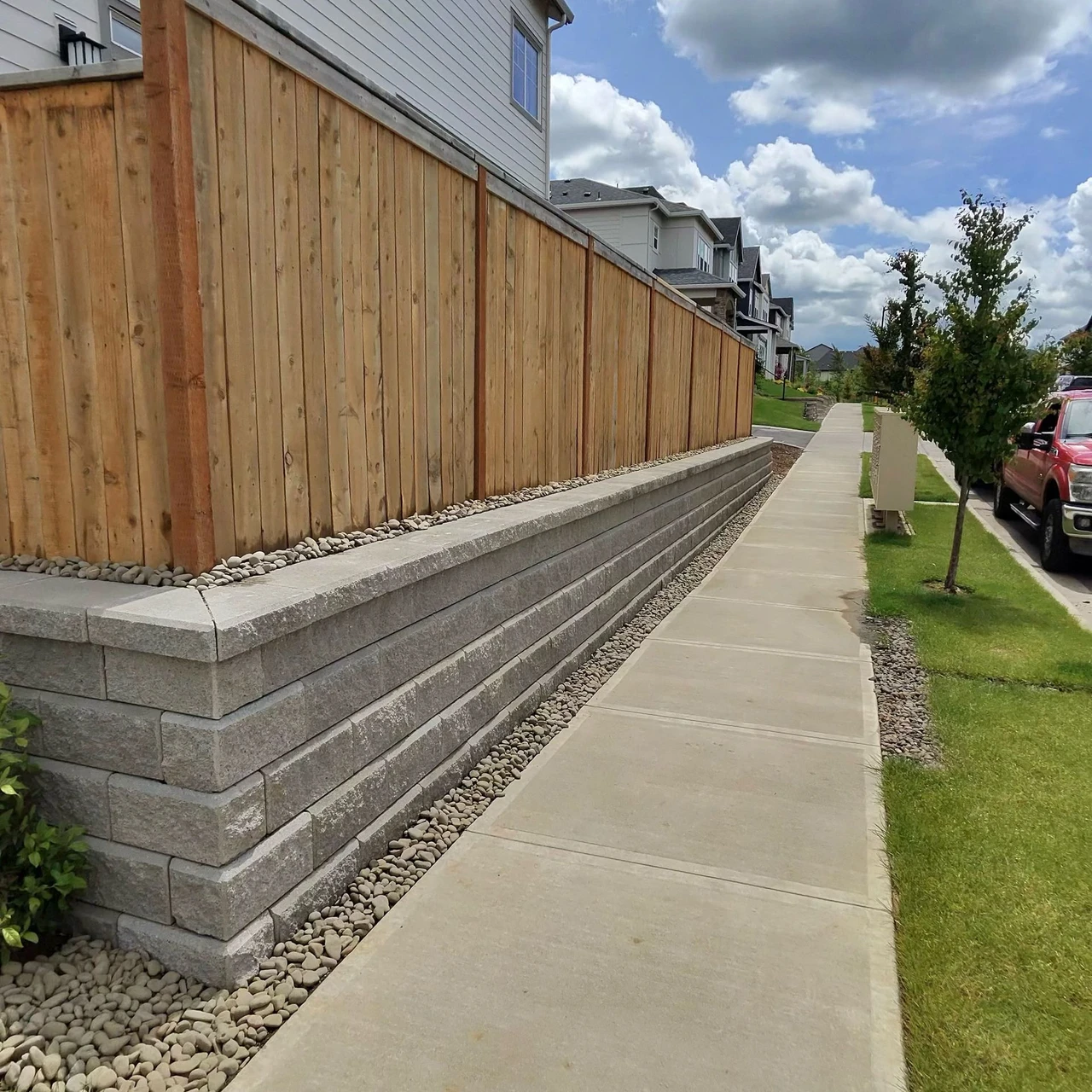 Wooden fence sitting on top of a retaining wall. Sidewalk is to the side of the retaining wall