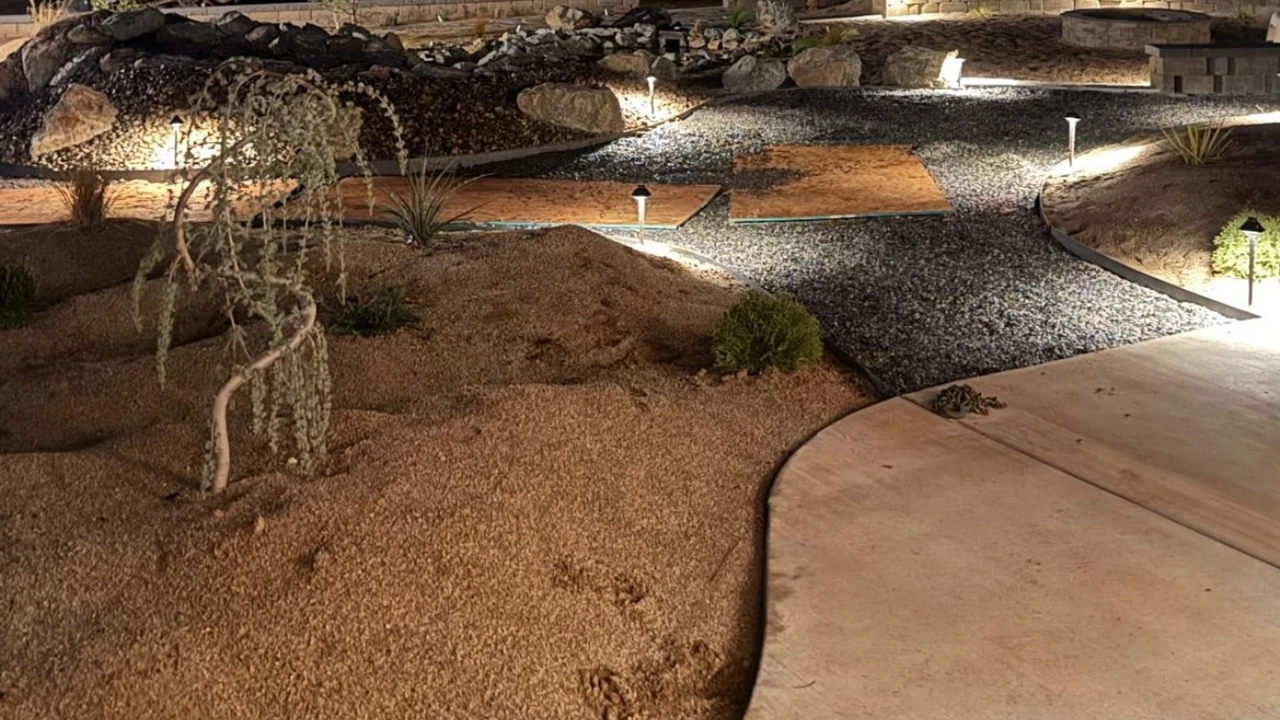 Nighttime view of a landscaped yard with curved concrete walkway, gravel and mulch beds, small shrubs and trees, low path lights, and a stone retaining wall under a dark sky.