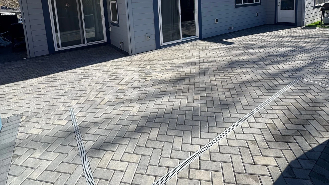 Gray interlocking paver patio in front of a modern house with sliding glass doors, windows, and dark trim under a clear blue sky.