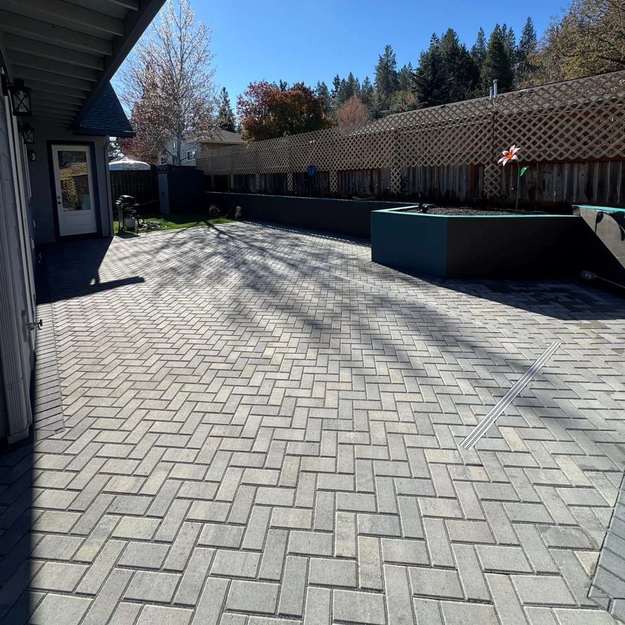 Sunny backyard patio with gray herringbone pavers, a raised planter box, house eaves along the left, a lattice-topped fence at the back, and trees beyond the yard.