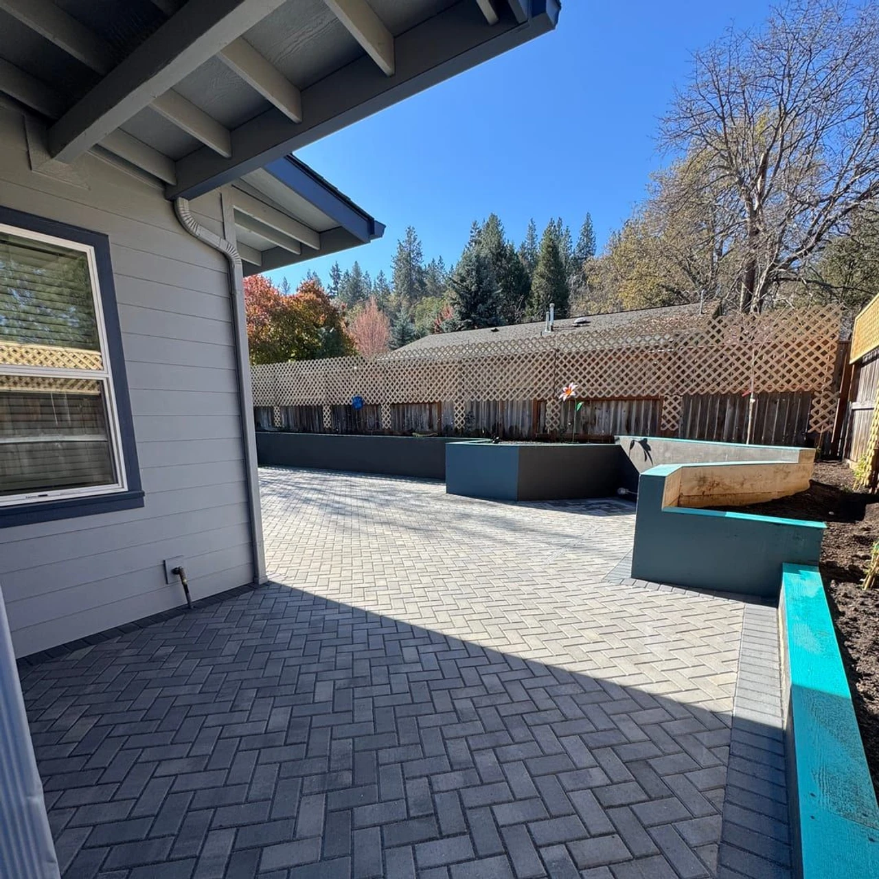 Residential backyard patio with gray herringbone pavers beside a house, raised planter walls, a wooden lattice fence, and trees under a clear blue sky.