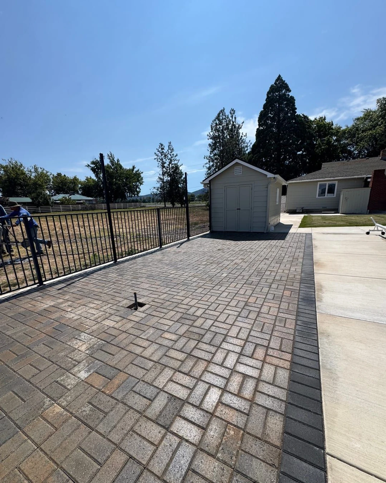 A residential backyard patio with gray brick pavers beside a concrete walkway, a small storage shed, black metal fence, house exterior, and trees under a clear blue sky.