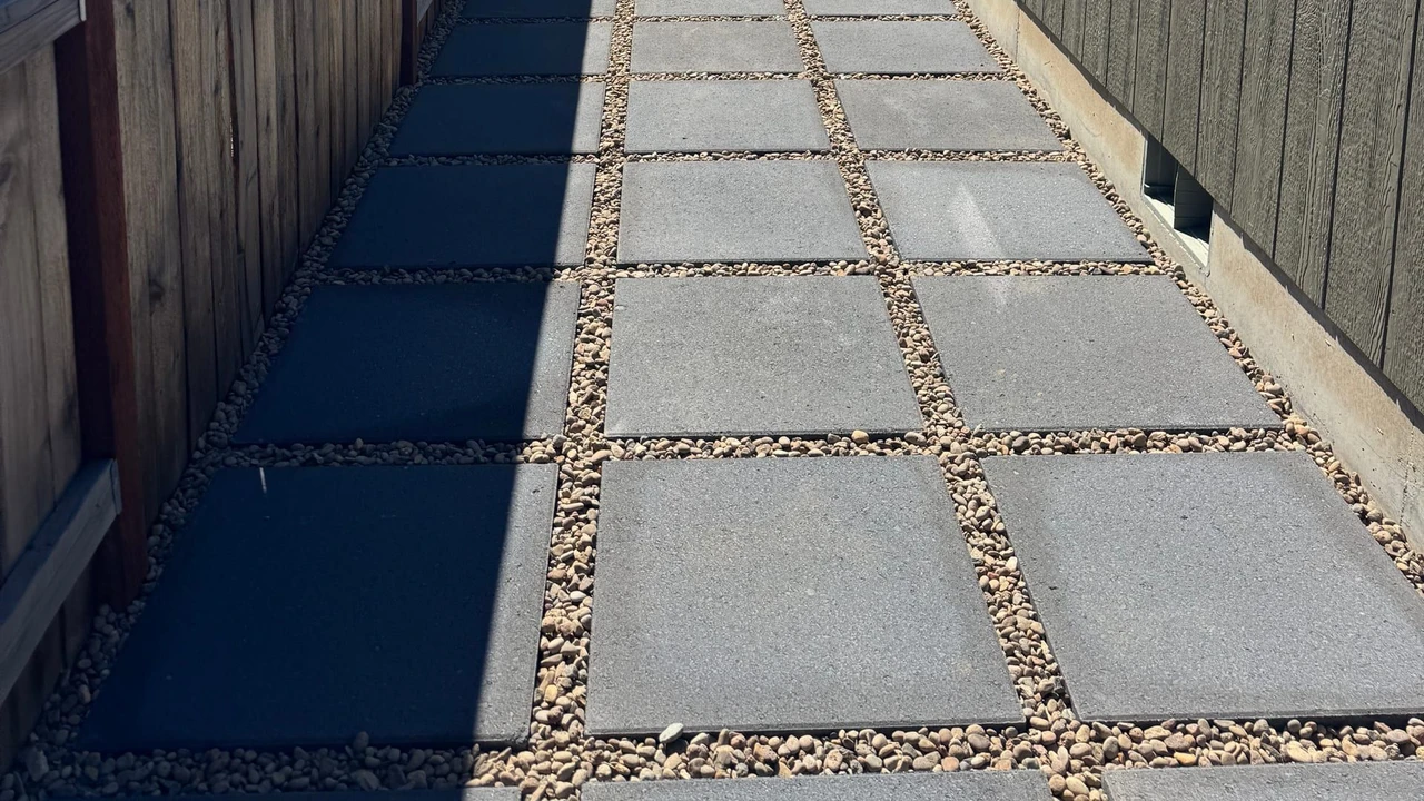 A narrow side-yard walkway with square concrete pavers set in gravel between a wooden fence and a dark house exterior, with windows, an air-conditioning unit, and blue sky overhead.