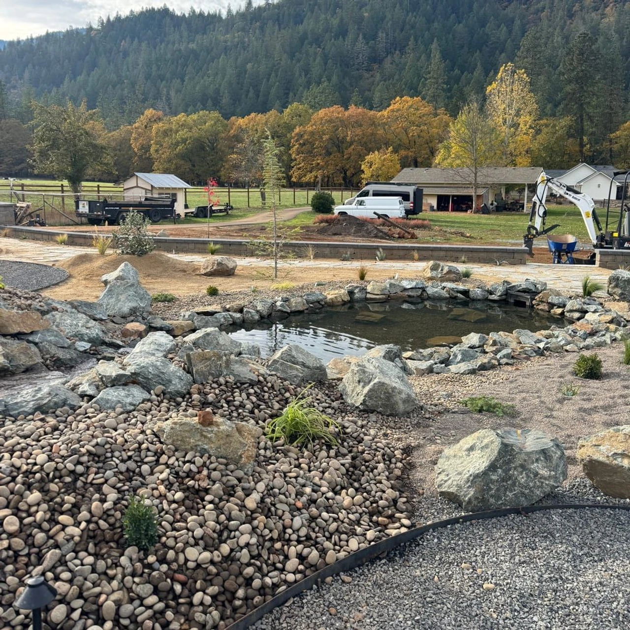 A landscaped yard with a rock-lined pond or water feature, gravel beds, large boulders, small plants, and a visible excavator. Trucks and a house sit in the background with forested hills and autumn trees beyond.