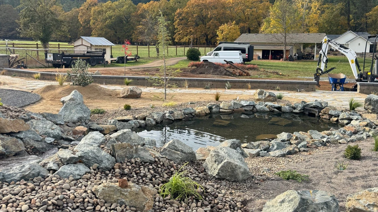 A landscaped yard with a rock-lined pond or water feature, gravel beds, large boulders, small plants, and a visible excavator. Trucks and a house sit in the background with forested hills and autumn trees beyond.