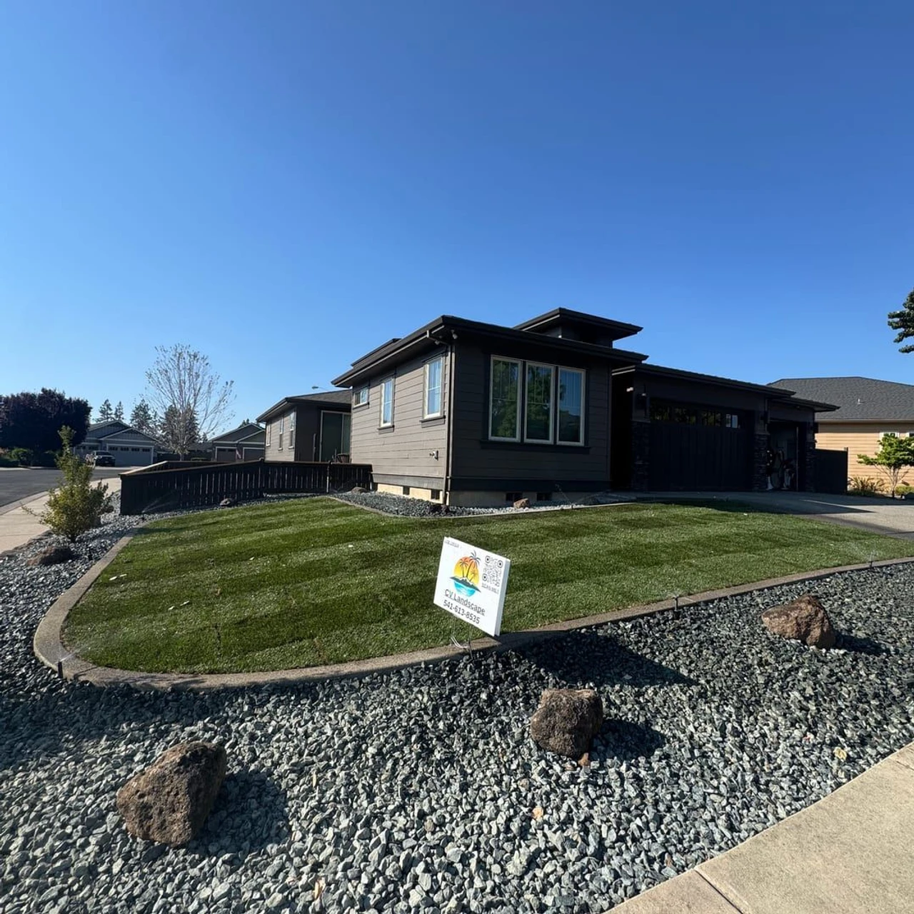 A newly landscaped suburban front yard with synthetic turf, gray rock ground cover, scattered decorative boulders, curved concrete edging, and a modern house under a clear blue sky.