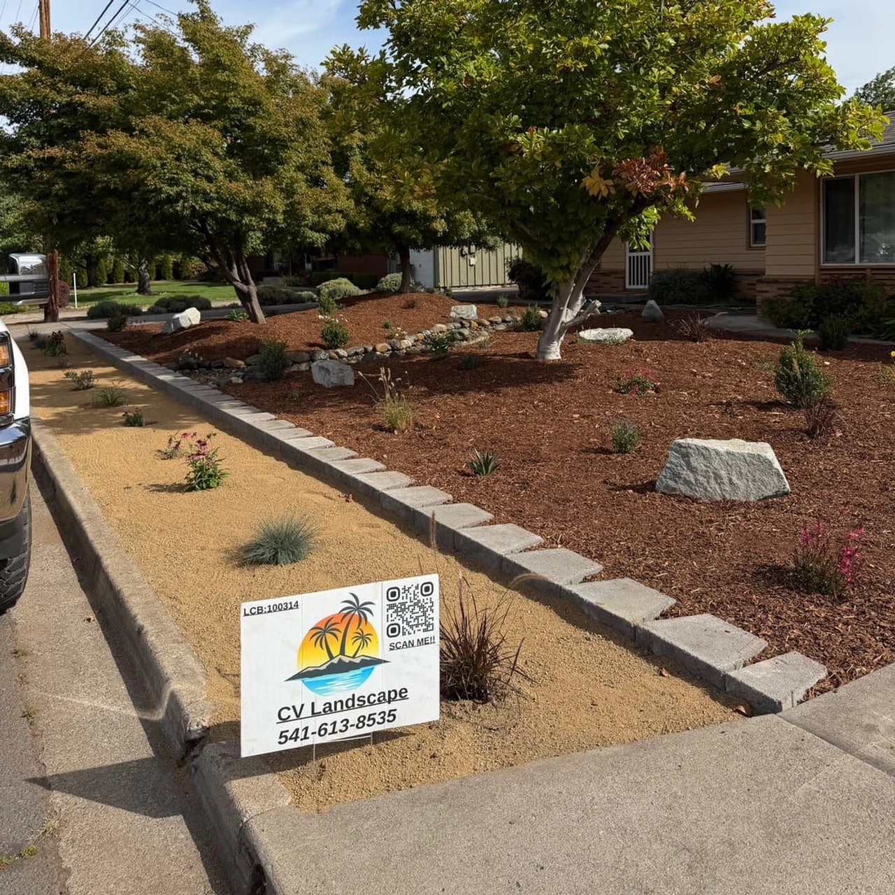 Residential front yard landscaping with gravel beds, small trees and shrubs, decorative rocks, a curb-edged strip by the sidewalk, and a lawn sign in the foreground.