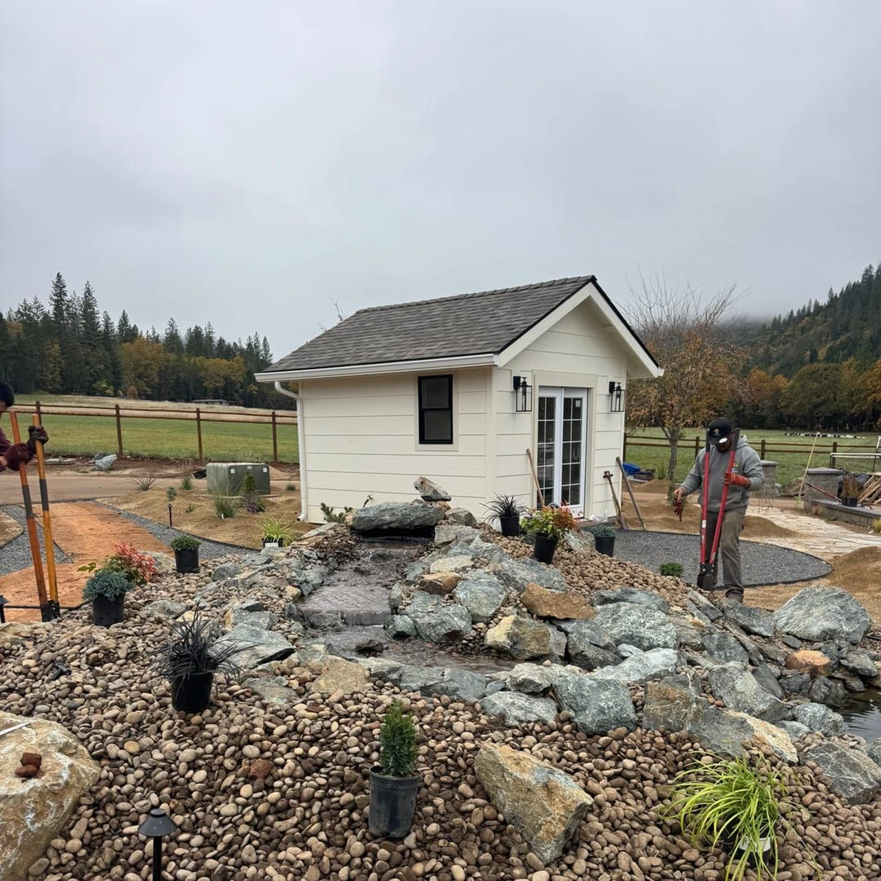 A landscaped yard with a small white shed, a rock waterfall or stream feature, rounded river stones, gravel edging, potted plants, and two workers using long-handled tools in a rural outdoor setting.