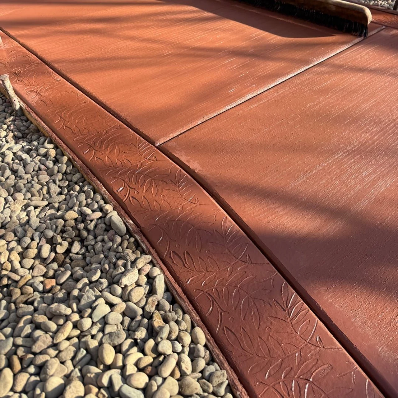 Close-up of a red-stained concrete patio or walkway with a stamped leaf-pattern border beside gray gravel, with a worker’s legs, broom, and bucket visible at the edge.