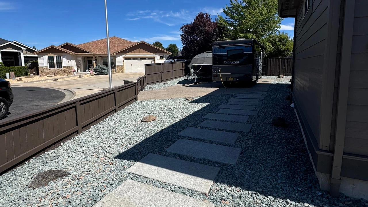 A residential side yard with large concrete stepping stones set in gray gravel beside a house, a brown fence, an RV or trailer in the distance, neighboring homes, and a bright blue sky with wispy clouds.