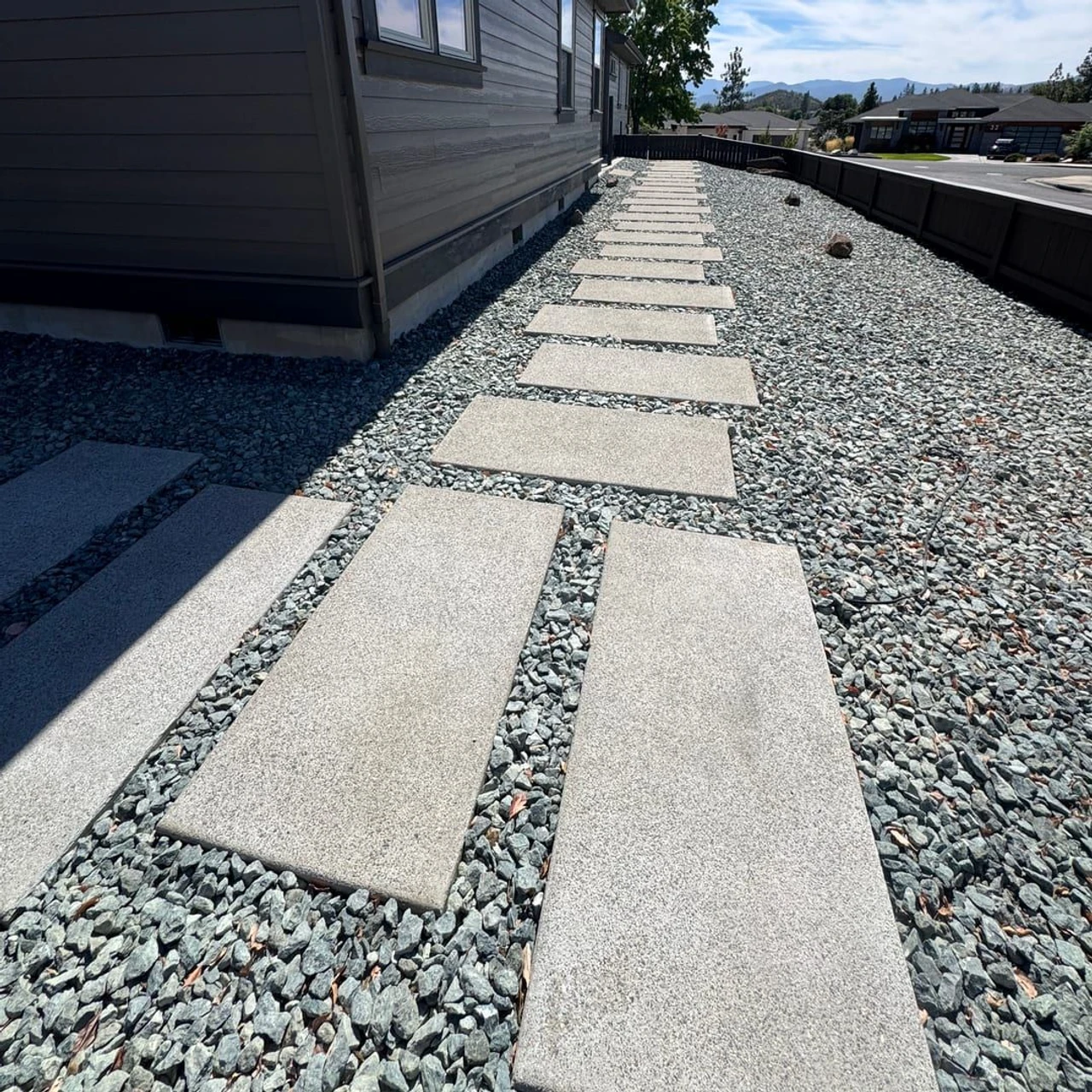 A residential side yard with large rectangular stepping stones set in gray gravel beside a house, bordered by a dark fence, with blue sky and distant houses in the background.