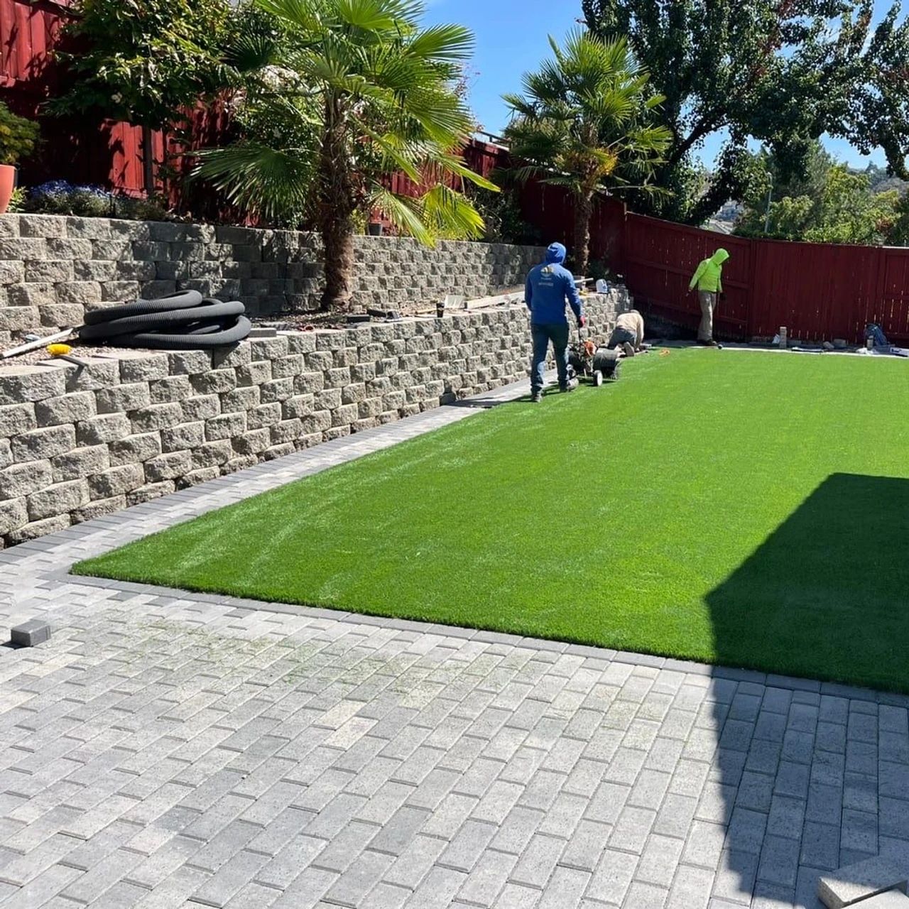 Backyard landscaping work with bright green synthetic turf bordered by gray pavers, stone retaining walls, red fence, palm trees, and three workers using tools under a blue sky.