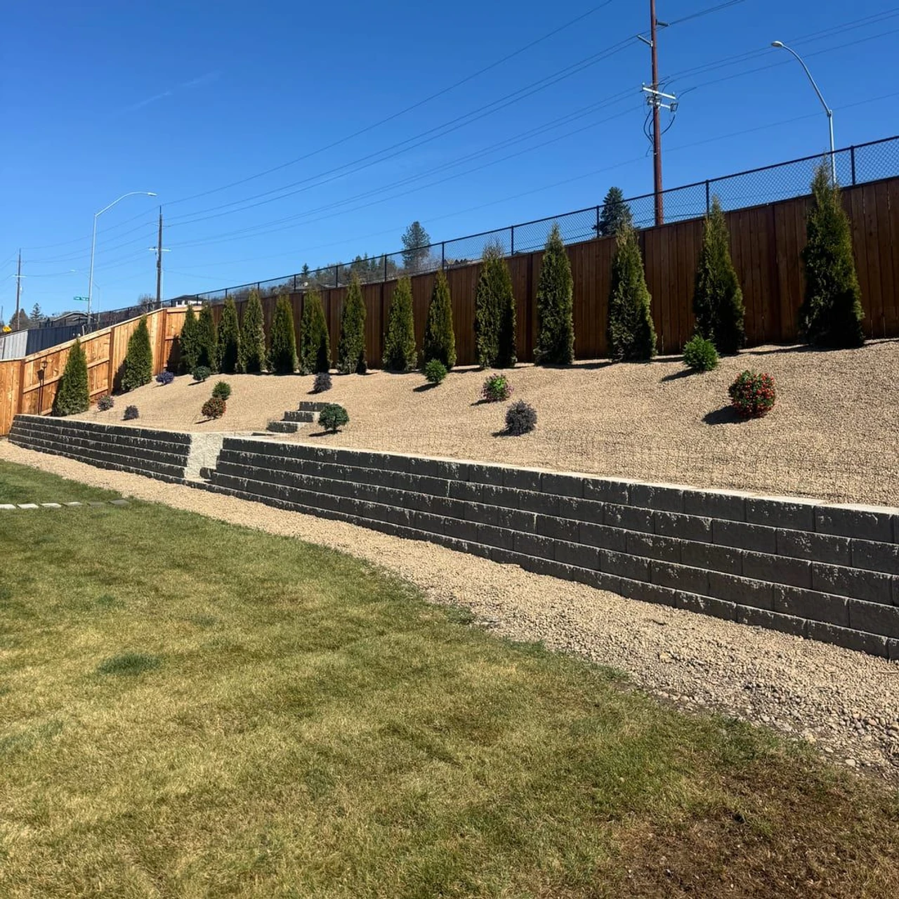 A landscaped sloped yard with dark retaining walls, gravel ground cover, rows of small conifer trees, a wooden fence, and a grassy lawn under a clear blue sky.